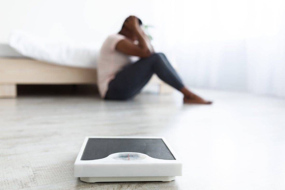 Person sitting on the floor with head in hands beside a bathroom scale
