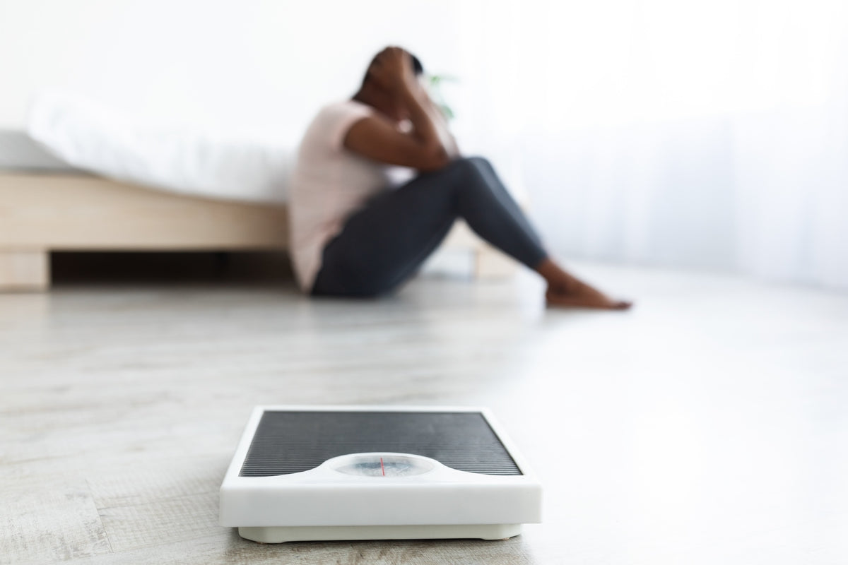 Person sitting on the floor with head in hands beside a bathroom scale