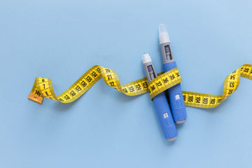Two GLP-1 injectors held together by measuring tape on a blue background, part of a discussion on what is GLP-1 diet.