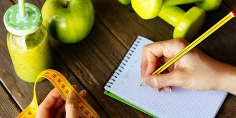 Person writing in a notebook beside a green smoothie and fresh apples