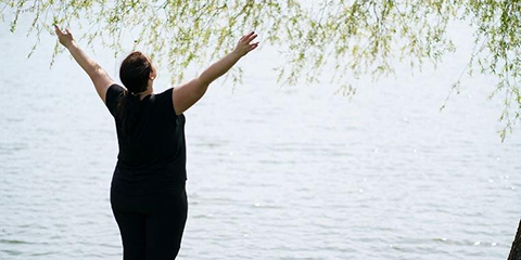 Person standing by a lake with arms raised toward the sky