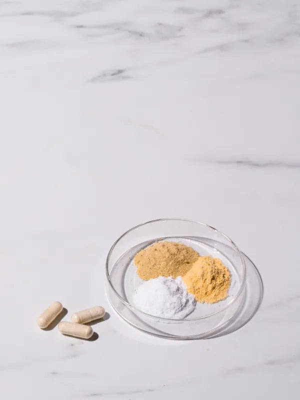 Four beige capsules and a small clear glass dish containing two piles of yellow powder and one pile of white powder, arranged on a white marble surface.